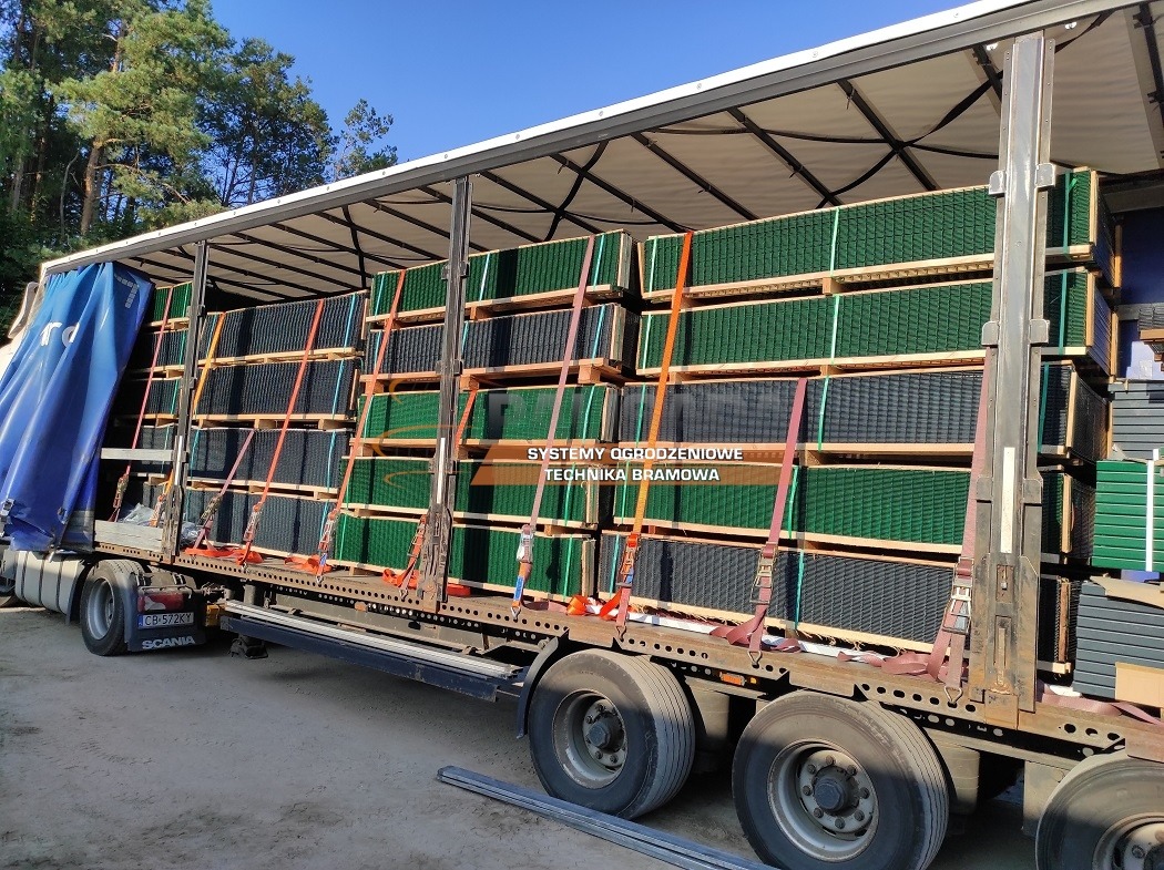 Transport paneli ogrodzeniowych na paletach, zabezpieczonych pasami, na naczepie ciężarówki Scania z niebieską plandeką w słoneczny dzień.
