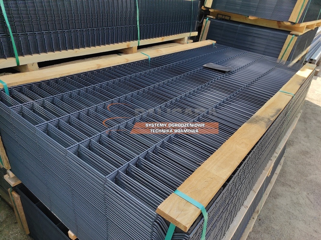 Stos paneli ogrodzeniowych z drutu zgrzewanego, zabezpieczonych drewnianymi belkami i taśmami transportowymi.