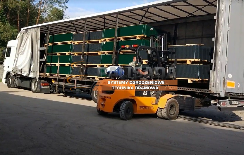 Pomarańczowy wózek widłowy ładuje palety z panelami ogrodzeniowymi w kolorze ciemnej zieleni do naczepy ciężarówki.