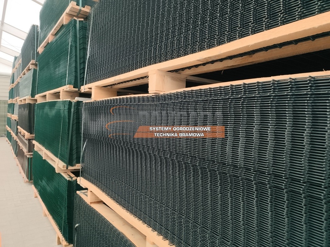 Stos paneli ogrodzeniowych zgrzewanych, w kolorze zielonym i ciemnoszarym, ułożonych na drewnianych paletach w magazynie.