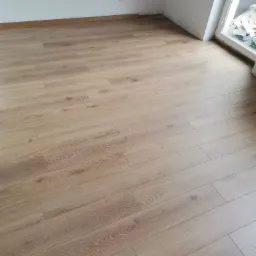 Jasne panele podłogowe ułożone w nowym pokoju z białymi ścianami i oknem dachowym, widoczne szczeliny dylatacyjne przy ścianach.