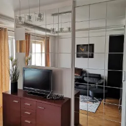 Salon w Piastowie z lustrzaną ścianą z kwadratowych paneli, odbijającą wnętrze z czarną sk&oacute;rzaną sofą i geometrycznym dywanem, brązowa komoda z telewizorem i dekoracyjną rośliną.