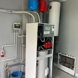 Nowoczesna kotłownia z białym piecem, zbiornikiem na wodę, niebieskim i czerwonym naczyniem przeponowym, rozdzielaczem ogrzewania podłogowego i niebieskim wiadrem na podłodze z szarych płytek.