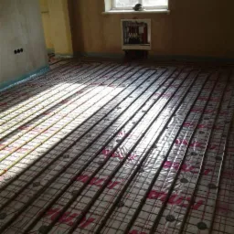Rozłożona instalacja ogrzewania podłogowego przed wylewką, widoczne rury grzewcze ułożone na folii z rastrem, szafka rozdzielcza na ścianie, surowy stan wykończenia ścian.