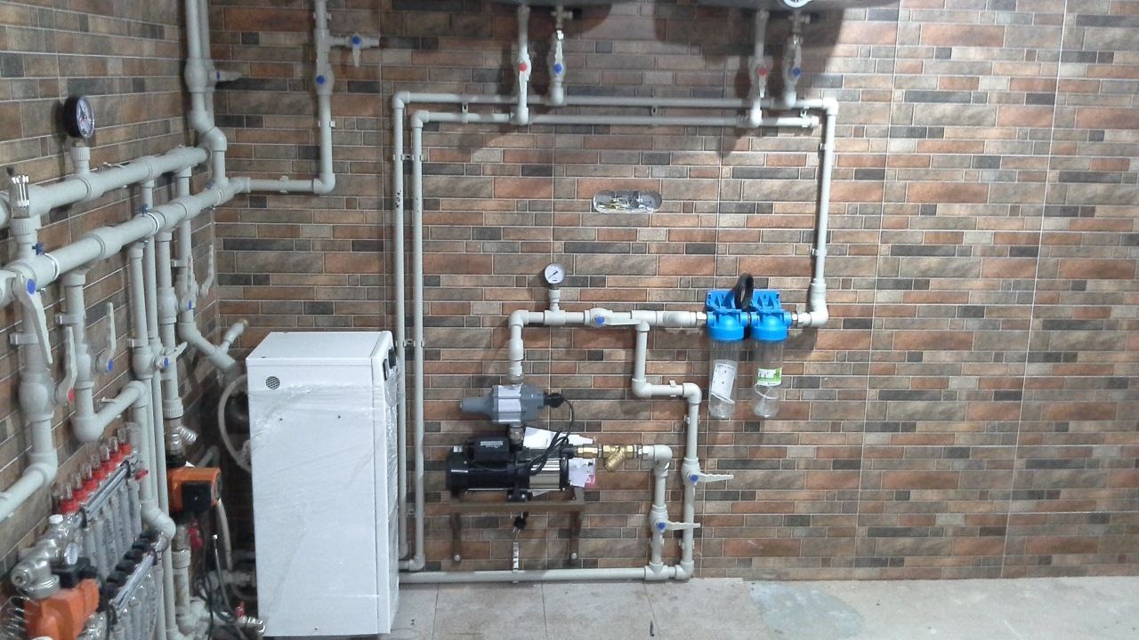 Złożony system rur, zaworów, manometrów, filtrów i pomp zamontowany na ścianie z płytek imitujących cegłę, z widocznym kotłem grzewczym owiniętym folią ochronną.