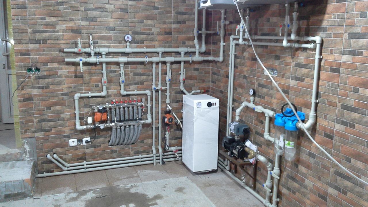 Rozbudowana instalacja centralnego ogrzewania z rozdzielaczem, pompami obiegowymi, filtrem wody i piecem w folii ochronnej, zamontowana na tle ściany wyłożonej płytkami imitującymi cegłę.