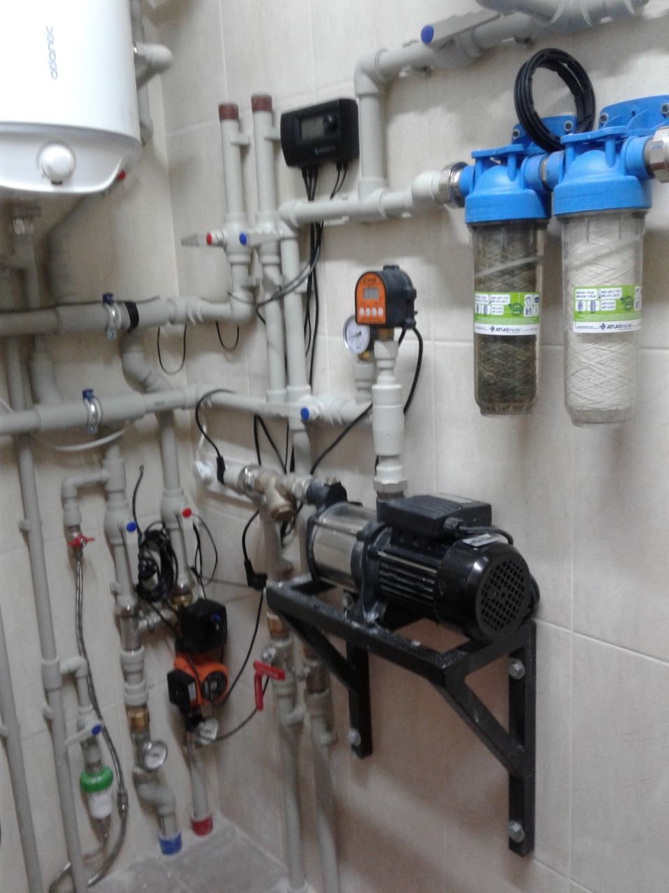 Pomieszczenie techniczne z zamontowanym bojlerem, pompą obiegową na czarnym wsporniku, filtrami wody z niebieskimi obudowami i licznymi rurami z tworzywa sztucznego.