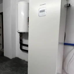 Biała pompa ciepła LG Therma V, zamontowana w pomieszczeniu z szarą podłogą i białymi ścianami, obok biały zasobnik ciepłej wody użytkowej.