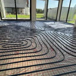 Niebieskie rury ogrzewania podłogowego ułożone spiralnie na folii z czerwoną siatką w jasnym pomieszczeniu z dużymi oknami, widok na zewnątrz.