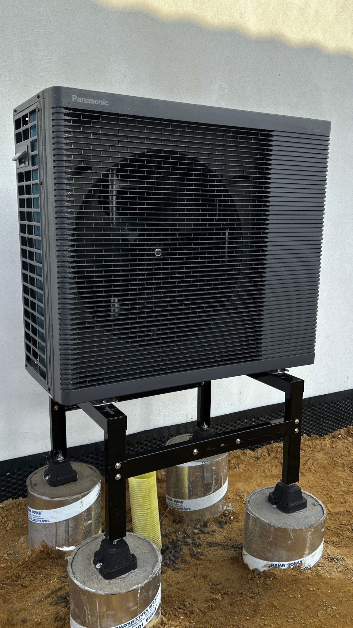 Czarna pompa ciepła Panasonic Aquarea zamontowana na metalowej konstrukcji, stojąca na betonowych fundamentach przy białej ścianie budynku. Widoczny żółty przewód.