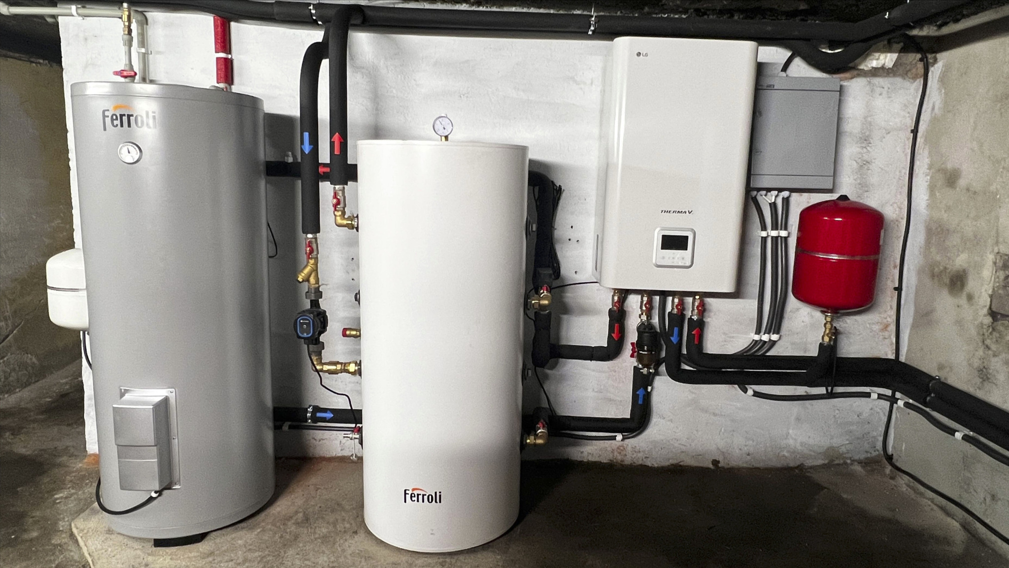 Dwa zbiorniki Ferroli, pompa ciepła LG Therma V i czerwony zbiornik wyrównawczy, połączone rurami w kotłowni z surowymi ścianami.