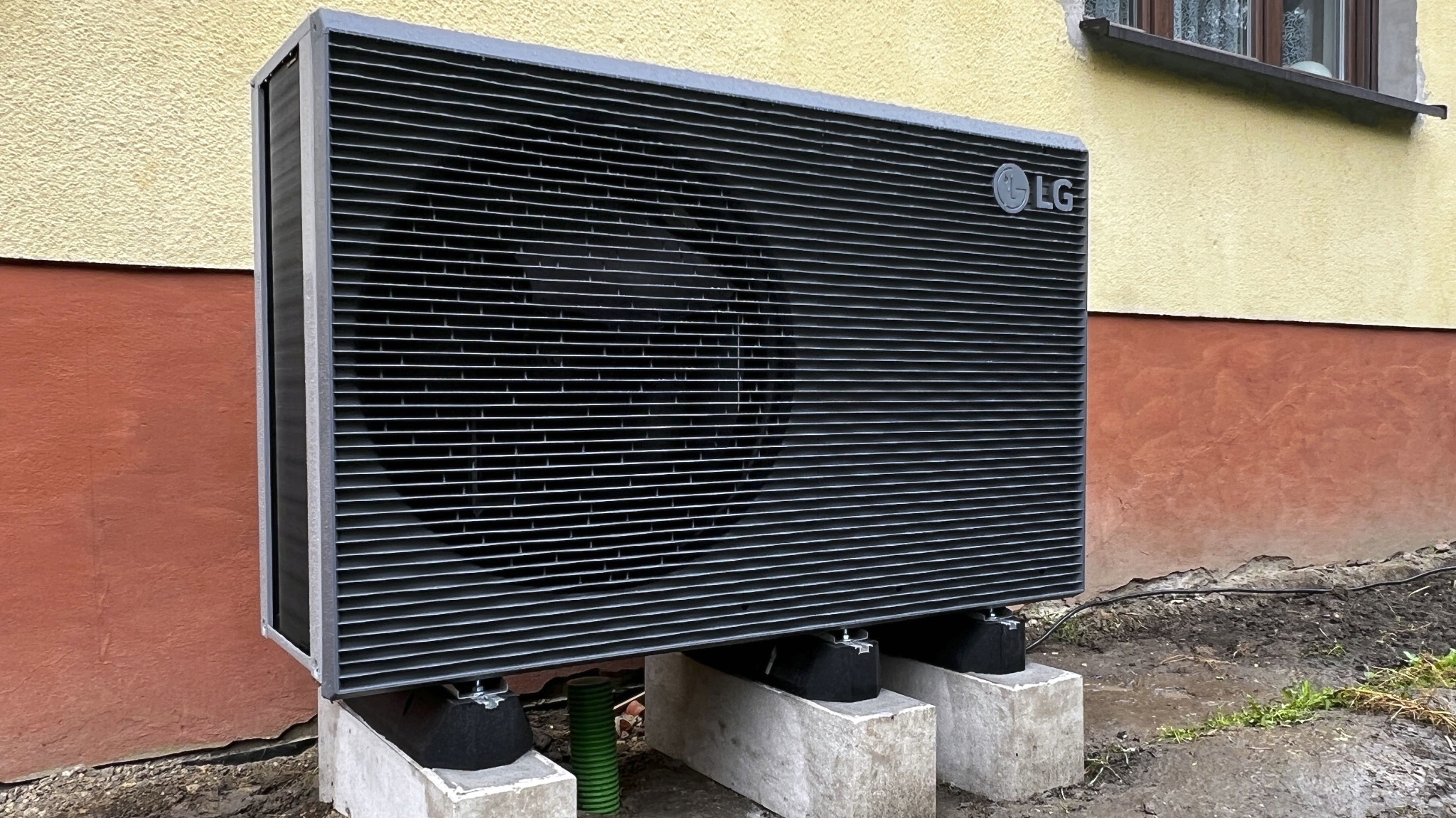 Zewnętrzna jednostka pompy ciepła LG, zamontowana na betonowych blokach przy ścianie budynku z widoczną instalacją. Ujęcie z bliska, podkreślające detale montażu.