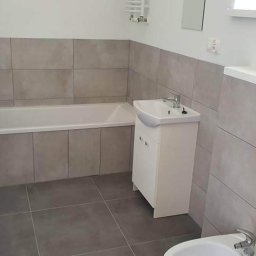 Remonty Wnętrz Poznań - Wyremontowana łazienka z szarymi płytkami, białą wanną i szafką pod umywalką oraz bidetem.