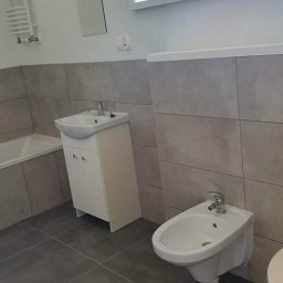 Remonty Wnętrz Poznań - Wykończona łazienka w Poznaniu z szarymi płytkami, białą umywalką z szafką, bidetem i wanną. Minimalistyczne lustro i grzejnik drabinkowy na ścianie.