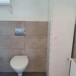 Świeżo wyremontowana toaleta z podwieszaną muszlą, szarą glazurą i białymi meblami łazienkowymi.