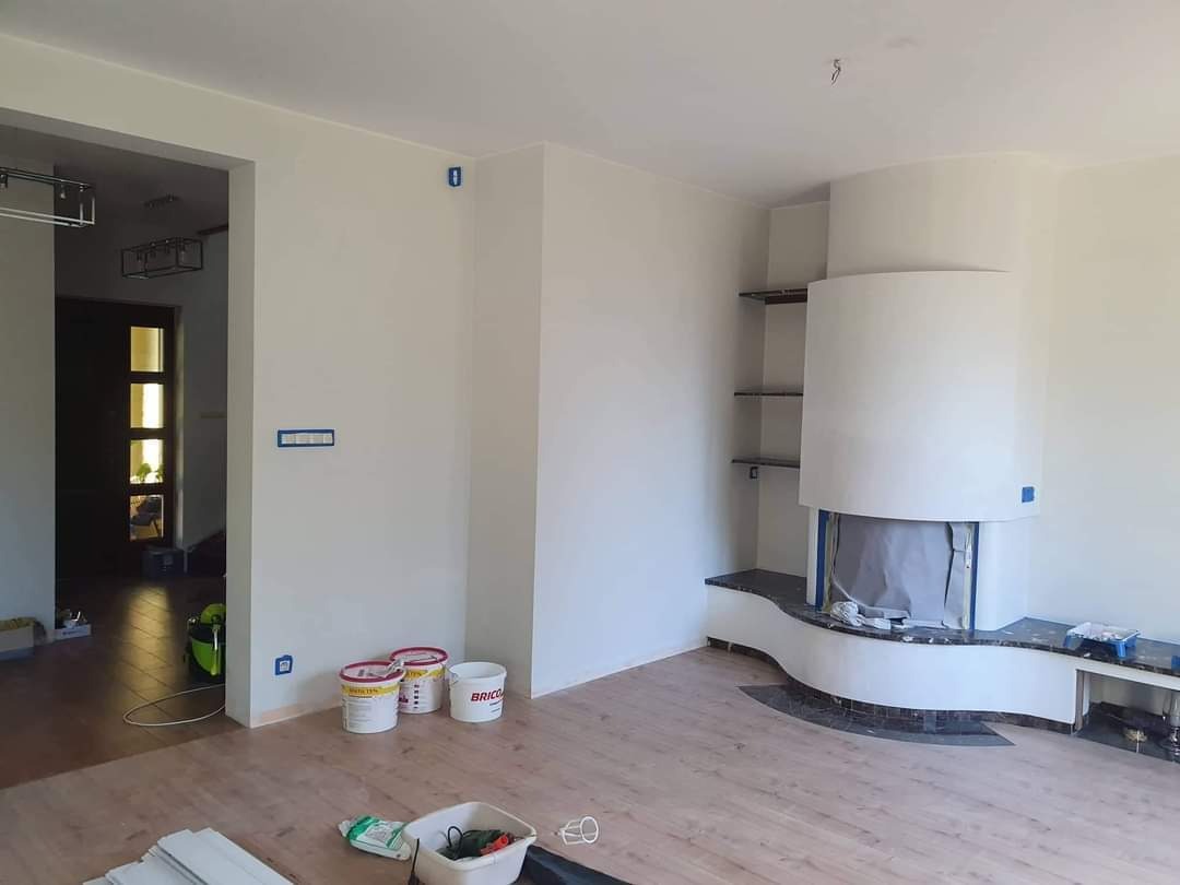 Wnętrze pokoju w trakcie malowania: ściany przygotowane z taśmą malarską, widoczne puszki z farbą, drewniana podłoga i obudowa kominka z półkami.