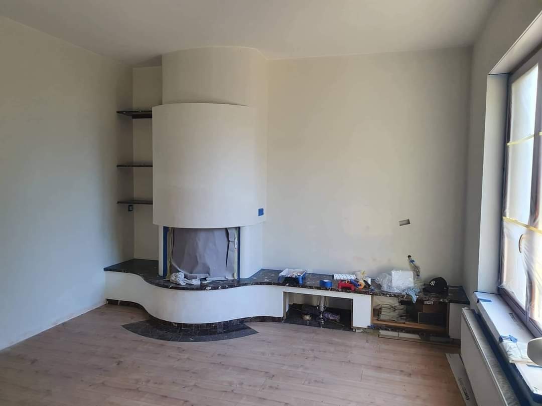 Pokój w trakcie prac wykończeniowych: ściany po malowaniu, zabezpieczone okno, zabudowany kominek z półkami, drewniana podłoga.