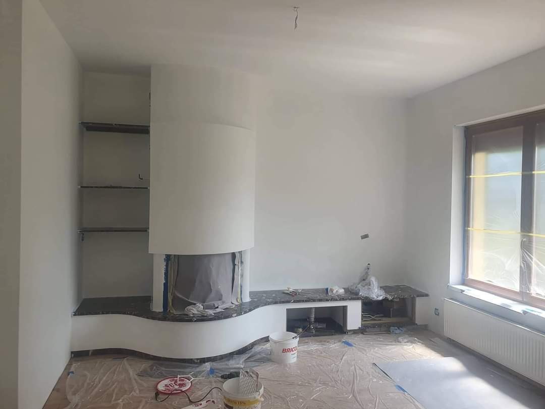 Pokój w trakcie malowania na biało, z widocznym kominkiem obłożonym czarnym kamieniem, zabezpieczonym folią i zarysowanymi półkami. Podłoga zabezpieczona folią malarską, widoczne puszki po farbie.