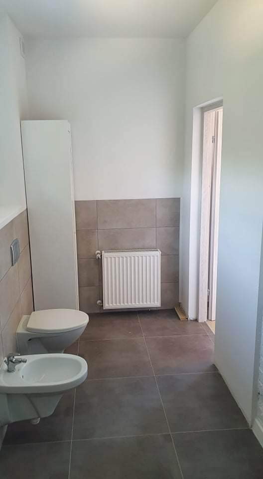 Jasna łazienka z szarymi płytkami, widoczne: toaleta, bidet, grzejnik i wysoka szafka