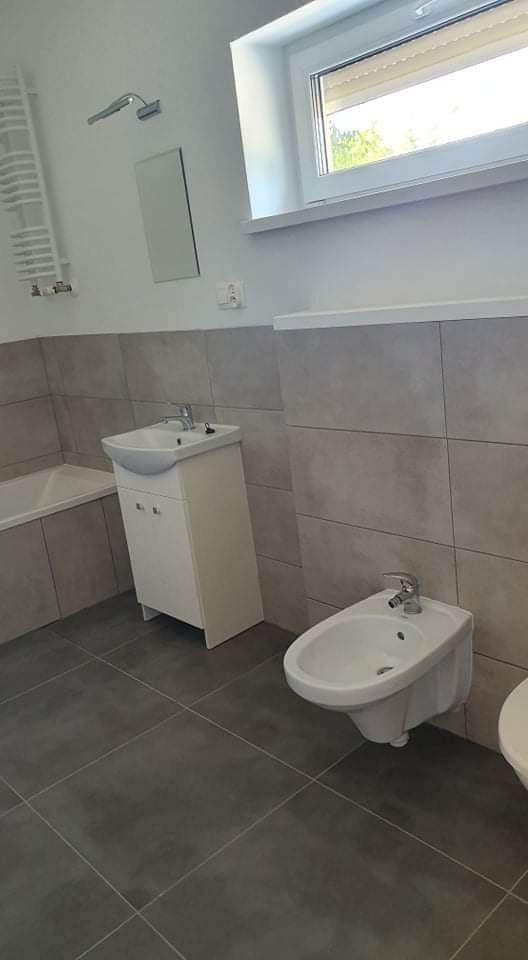 Wykończona łazienka w Poznaniu z szarymi płytkami, białą umywalką z szafką, bidetem i wanną. Minimalistyczne lustro i grzejnik drabinkowy na ścianie.