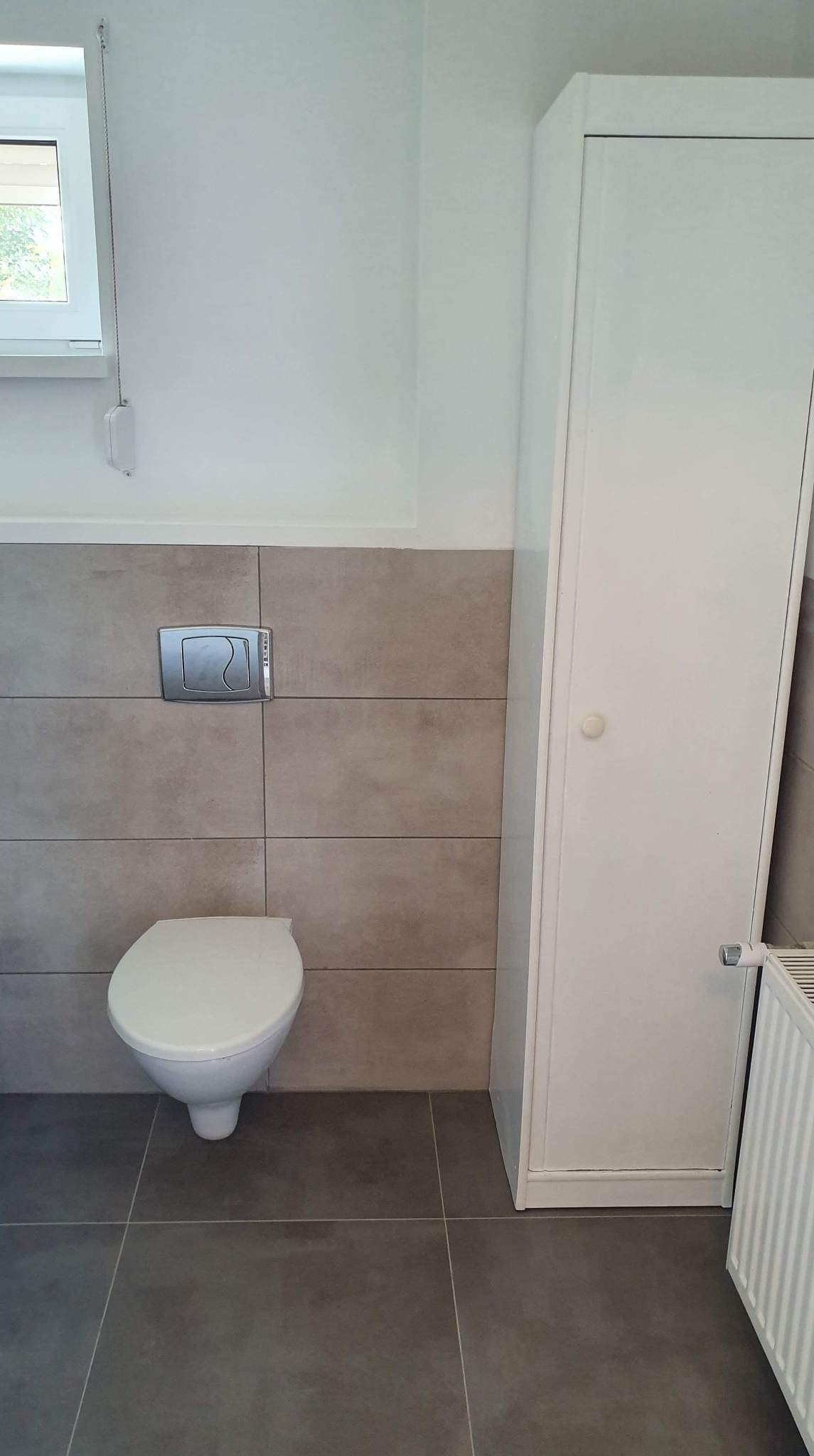 Świeżo wyremontowana toaleta z podwieszaną muszlą, szarą glazurą i białymi meblami łazienkowymi.