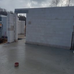 Zakład remontowo-budowlany Paweł Pastuszka - Wnętrze surowego domu w trakcie budowy, widoczne ściany z białych bloczków, wylany betonowy fundament, czerwona rura kanalizacyjna na podłodze, drzewa w tle.