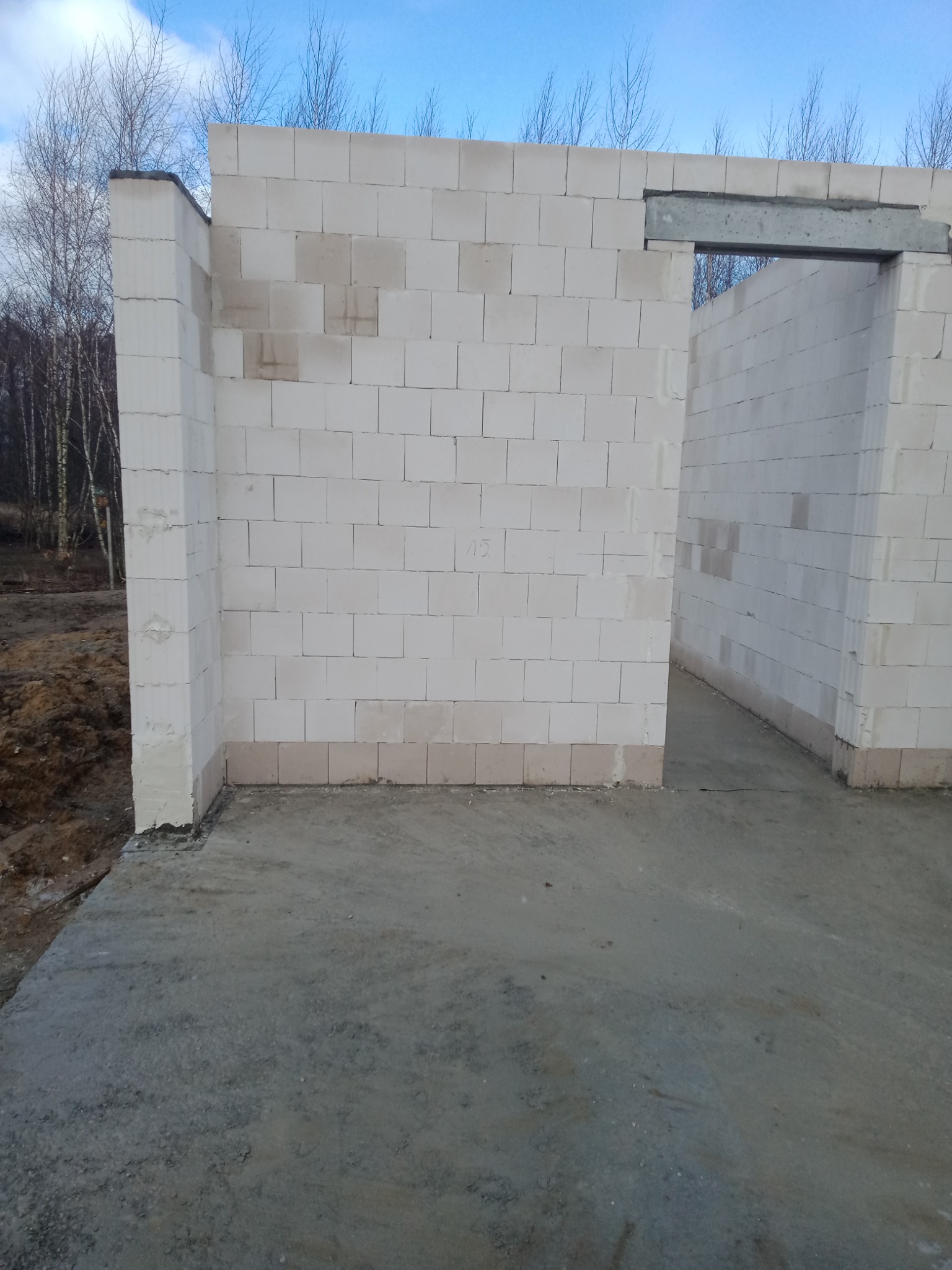 Wznoszone ściany z białych bloczków na fundamencie, widoczny otwór drzwiowy z betonowym nadprożem, w tle drzewa bez liści.