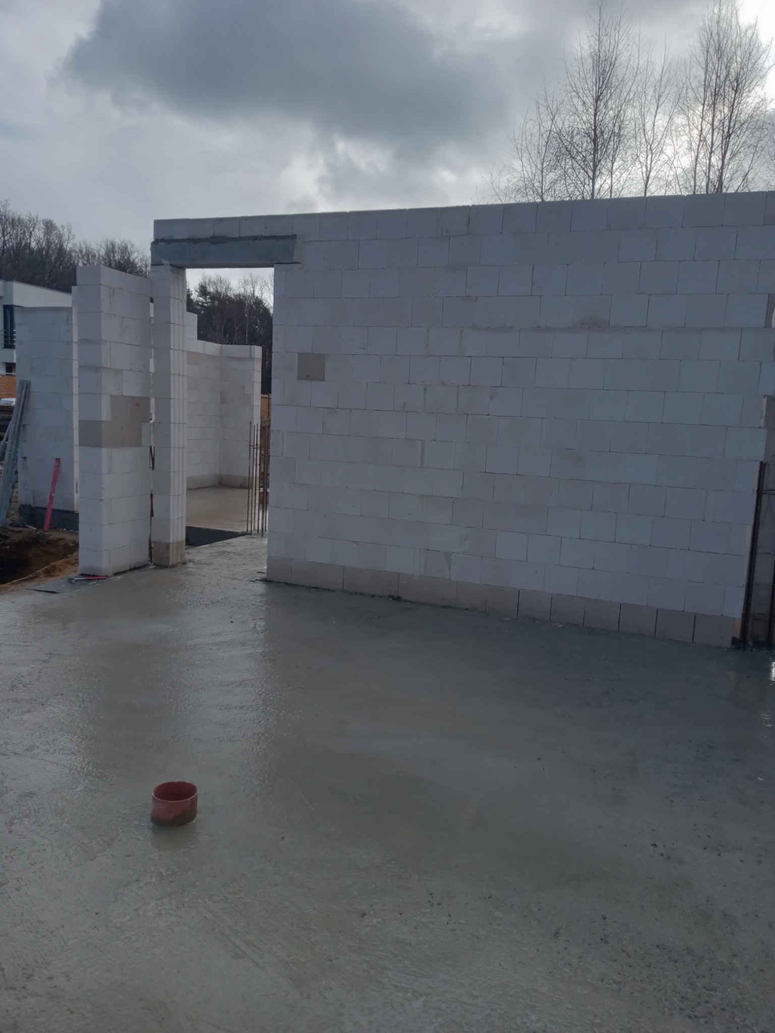 Wnętrze surowego domu w trakcie budowy, widoczne ściany z białych bloczków, wylany betonowy fundament, czerwona rura kanalizacyjna na podłodze, drzewa w tle.