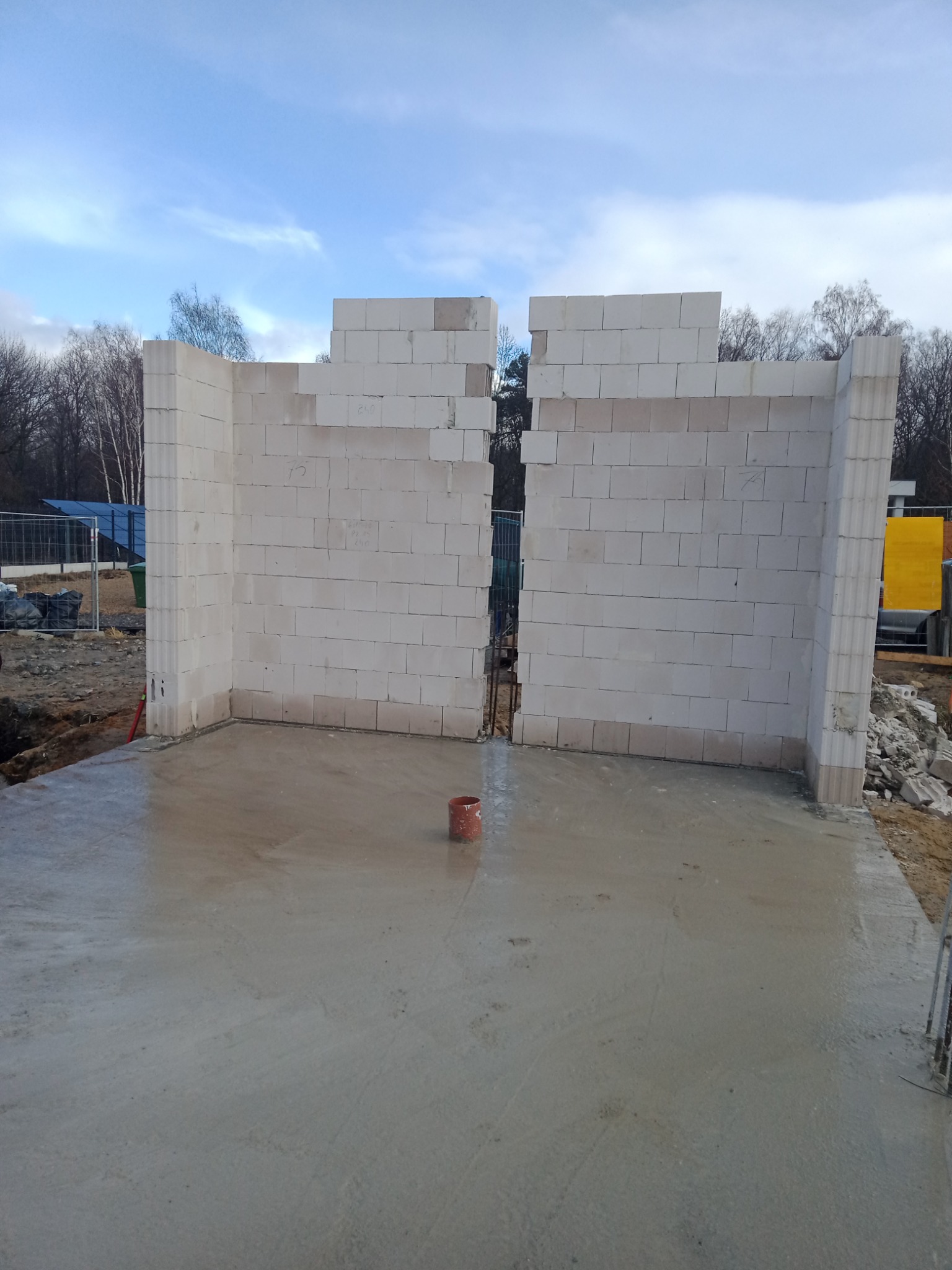 Wznoszone ściany z białych bloczków na wylewce fundamentowej, widoczny fragment instalacji oraz elementy rusztowania, drzewa w tle.