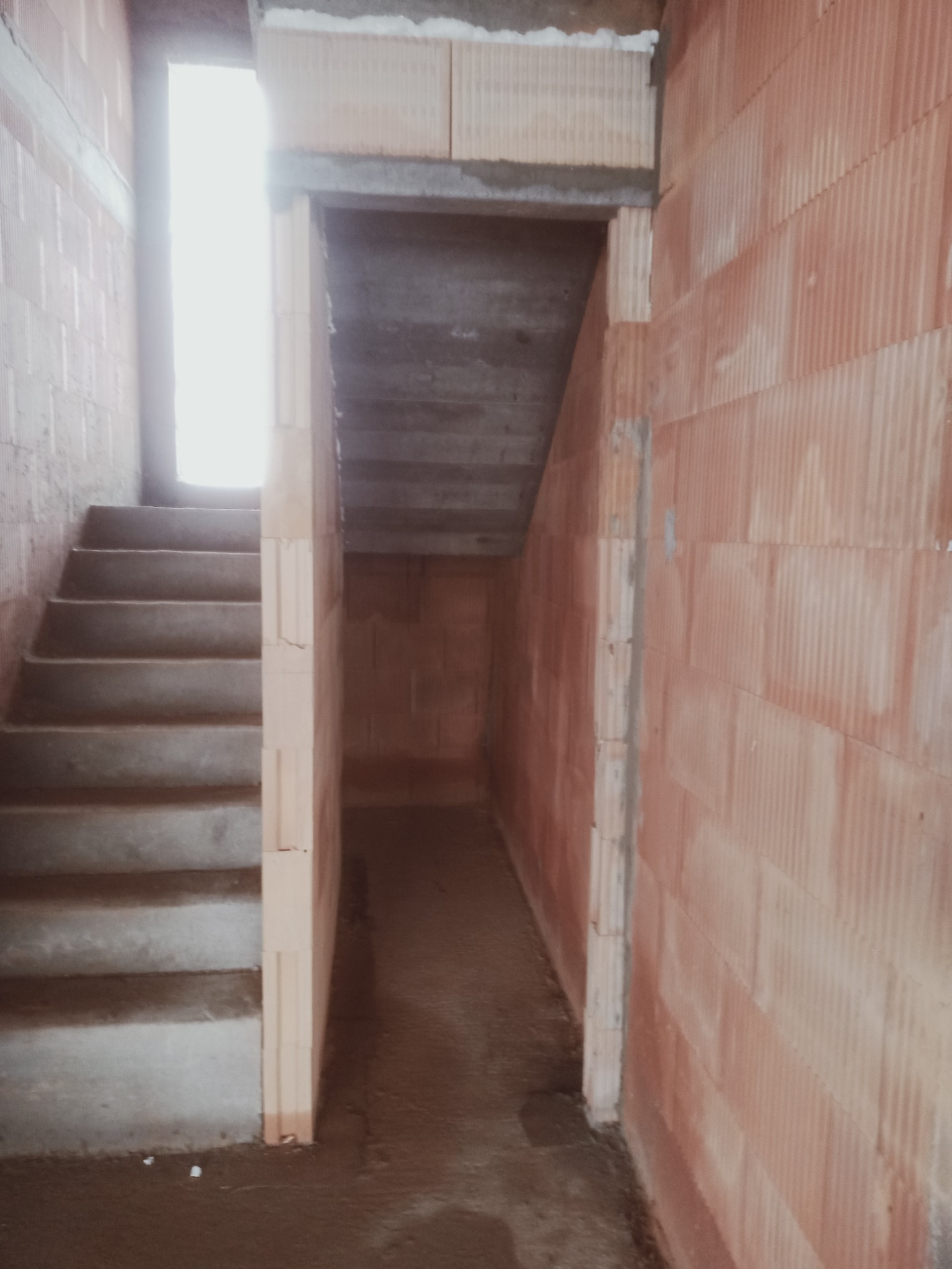 Wnętrze surowego domu w trakcie budowy: ściany z czerwonej cegły, betonowe schody prowadzące do góry i przejście do innego pomieszczenia.