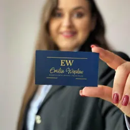 Kobieta w marynarce prezentuje wizytówkę w odcieniach granatu ze złotym napisem 'EW Emilia Więsław Kancelaria Radcy Prawnego', uśmiecha się w tle.