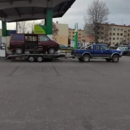 Niebieski pickup ciągnie przyczepę z uszkodzonym, bordowym vanem na stacji benzynowej BP, widoczne budynki mieszkalne w tle.