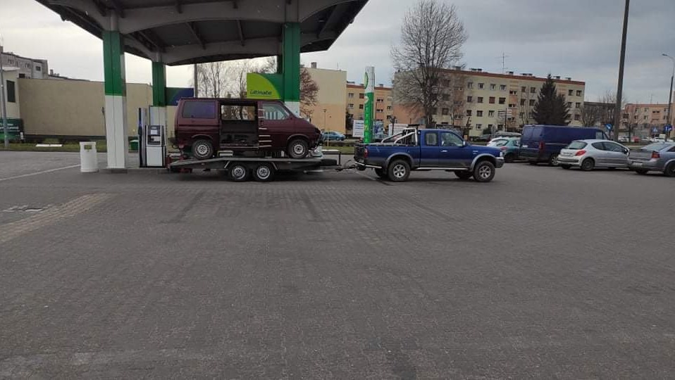 Niebieski pickup ciągnie przyczepę z uszkodzonym, bordowym vanem na stacji benzynowej BP, widoczne budynki mieszkalne w tle.