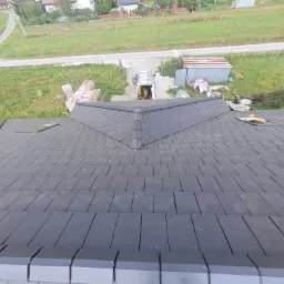 Widok z góry na nowy dach z ciemnej dachówki ceramicznej z zamontowanym oknem dachowym, w tle zaparkowany bus z drabiną i materiały budowlane.