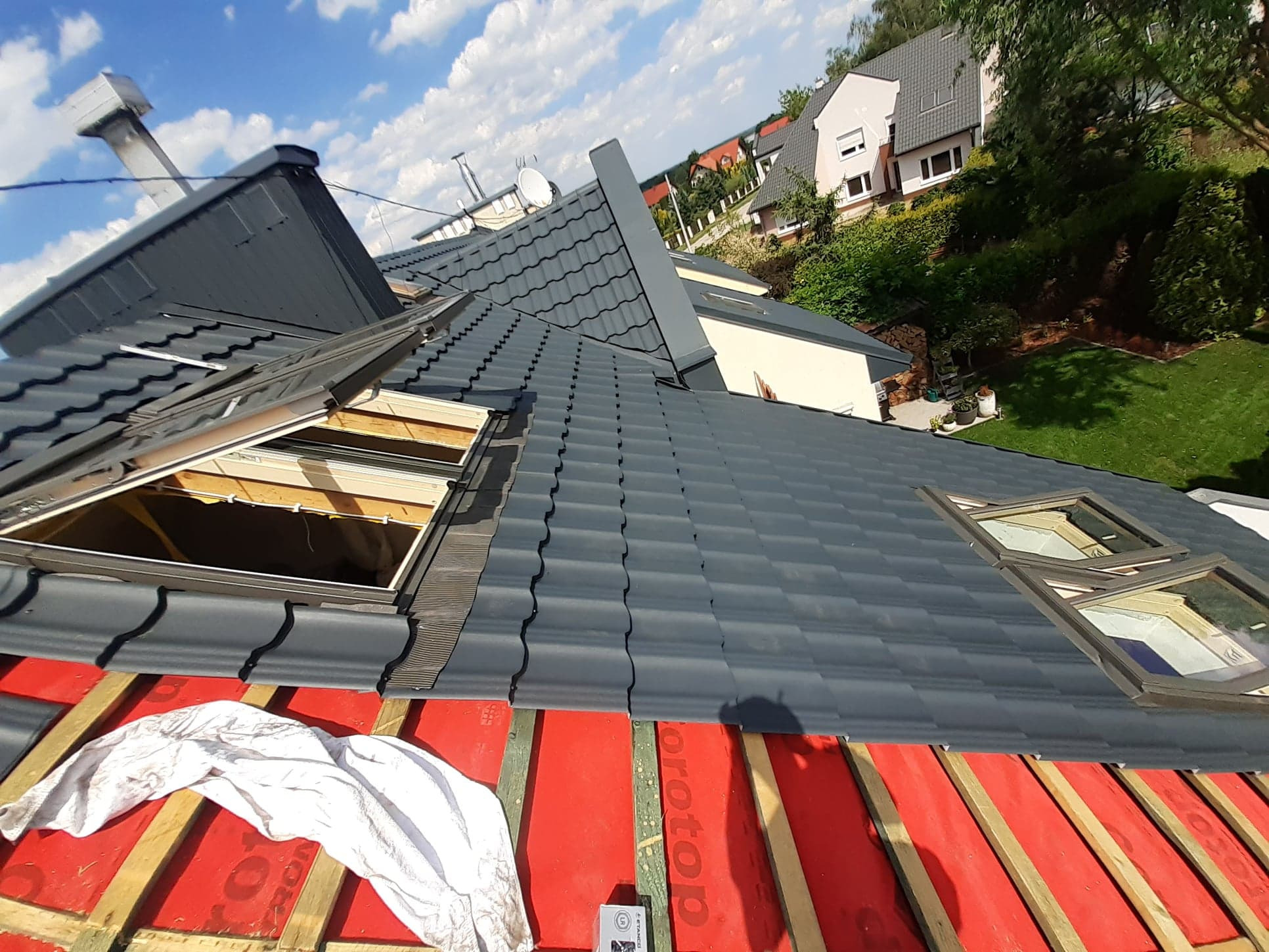 Szary dach z dachówki, częściowo pokryty czerwoną membraną, z otwartymi oknami dachowymi i białą szmatką na powierzchni, widok z góry na tle domów i zieleni.