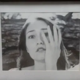 Oprawiona w ramę czarno-biała fotografia portretowa młodej kobiety, częściowo zasłaniającej twarz dłonią, z rozmytym tłem.