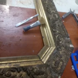 Fragment złoconej ramy z ornamentami, narzędzia konserwatorskie: pędzel i skalpel leżące na drewnianym blacie.