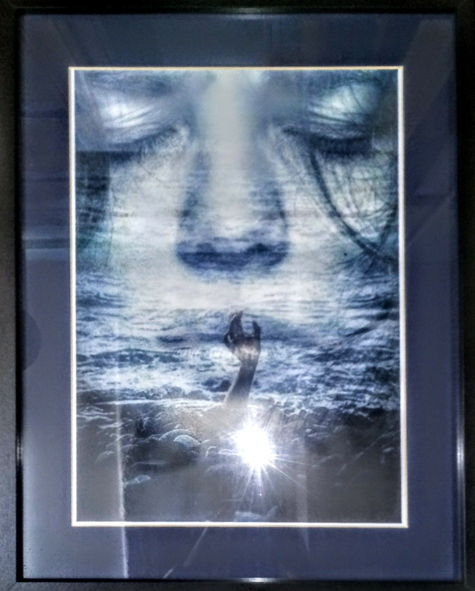 Oprawiony obraz przedstawiający twarz z zamkniętymi oczami nad powierzchnią wzburzonej wody, z wyciągniętą ręką w kierunku światła.