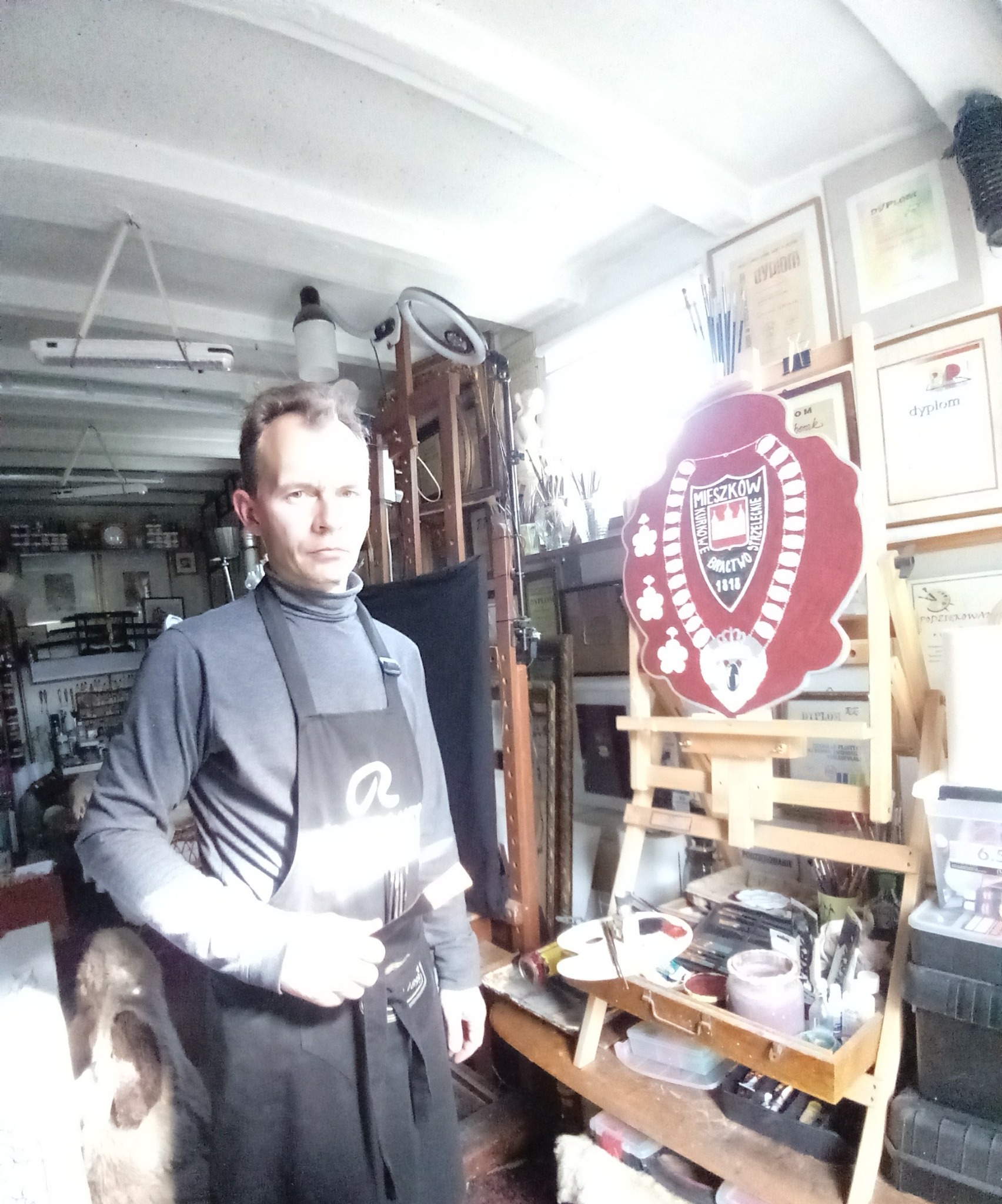 Artysta w pracowni, ubrany w czarny fartuch, na tle sztalugi z herbem, pędzli, farb i dyplomów na ścianie.