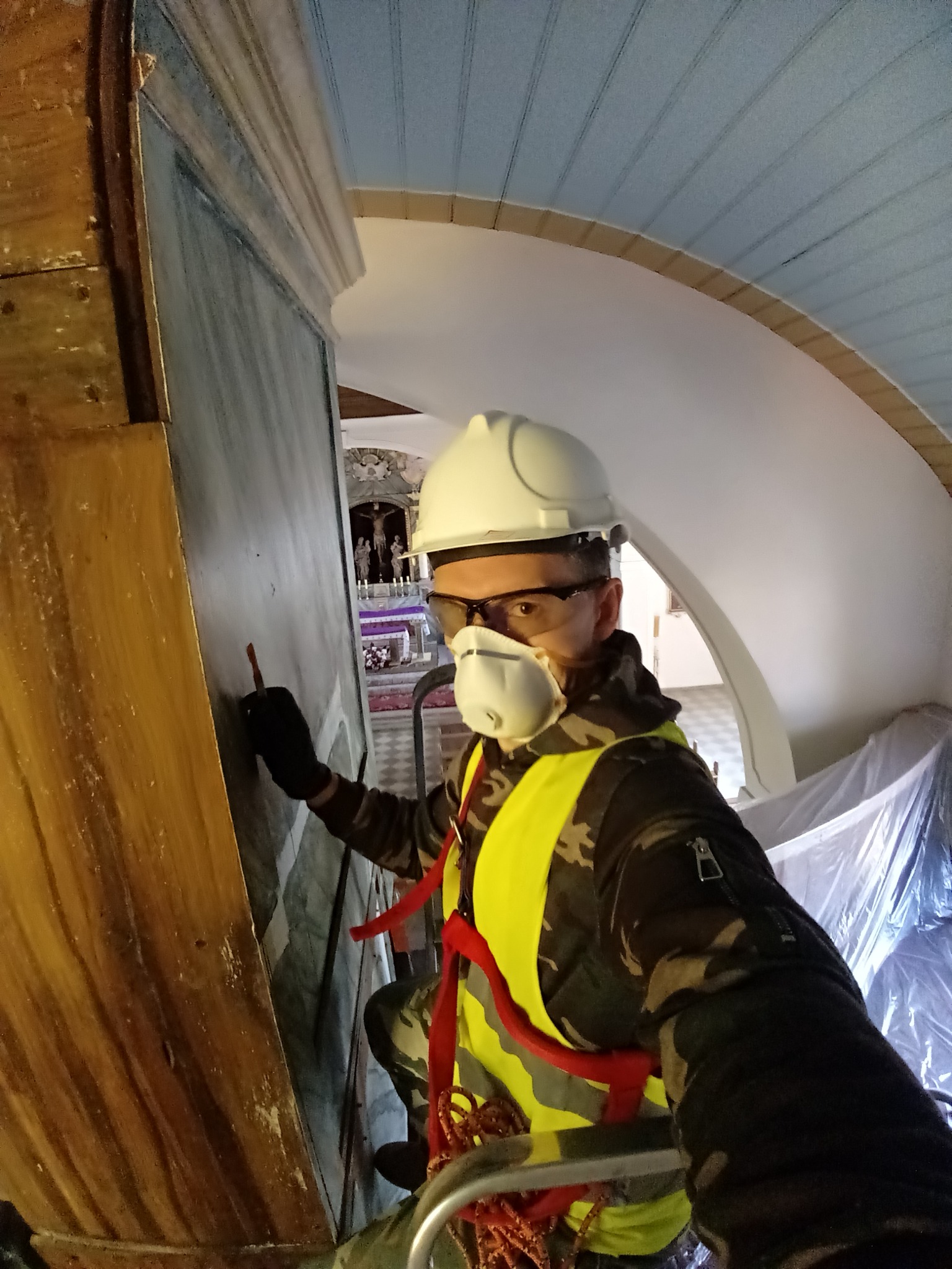 Pracownik w kasku i masce ochronnej, zabezpieczony linami, retuszuje malowidło na ścianie w kościele, używając małego pędzla.