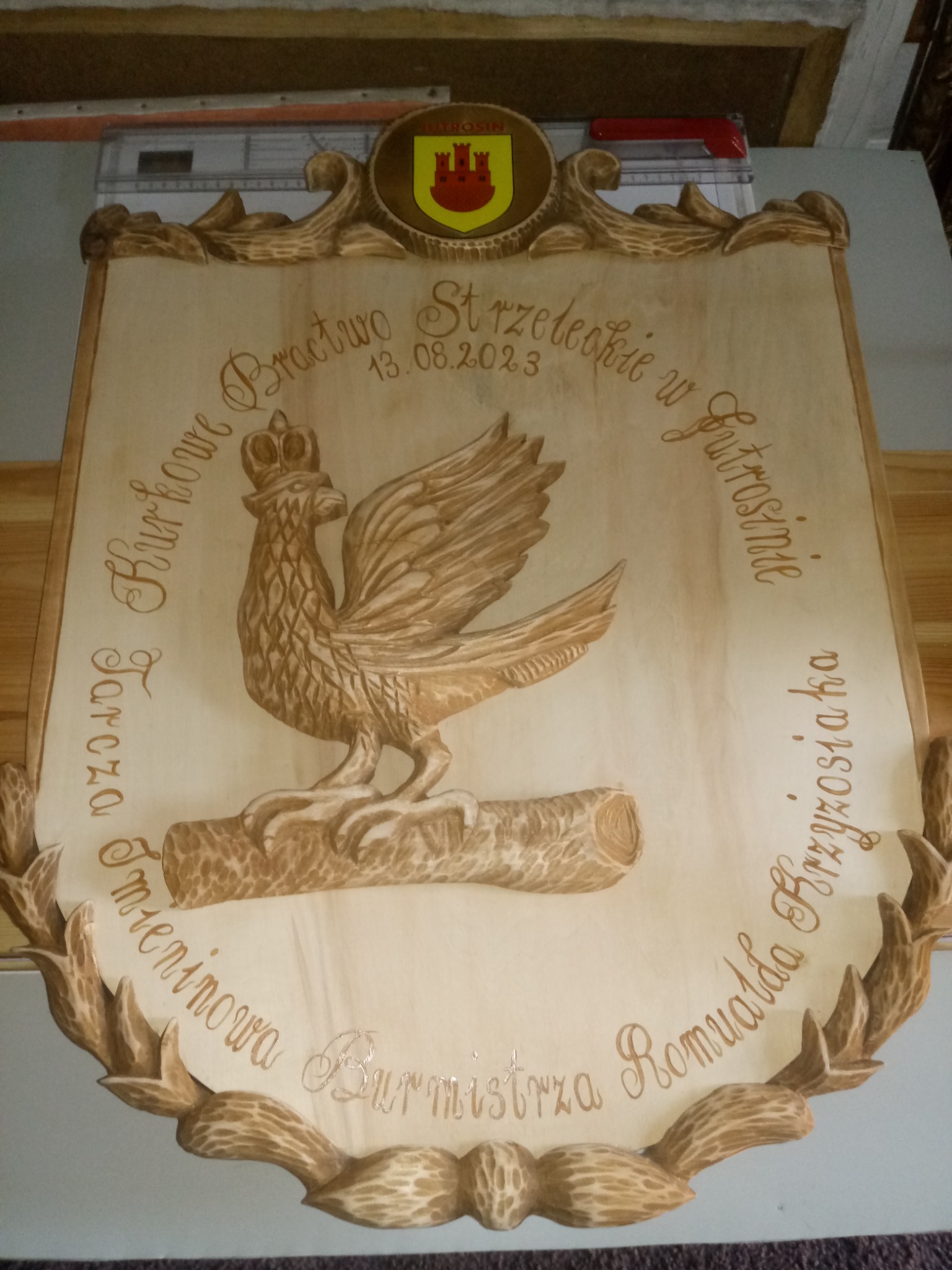 Drewniana tablica z płaskorzeźbą orła w koronie, otoczona wygrawerowanym napisem i dekoracyjną ramą z motywem liści, zwieńczona herbem.