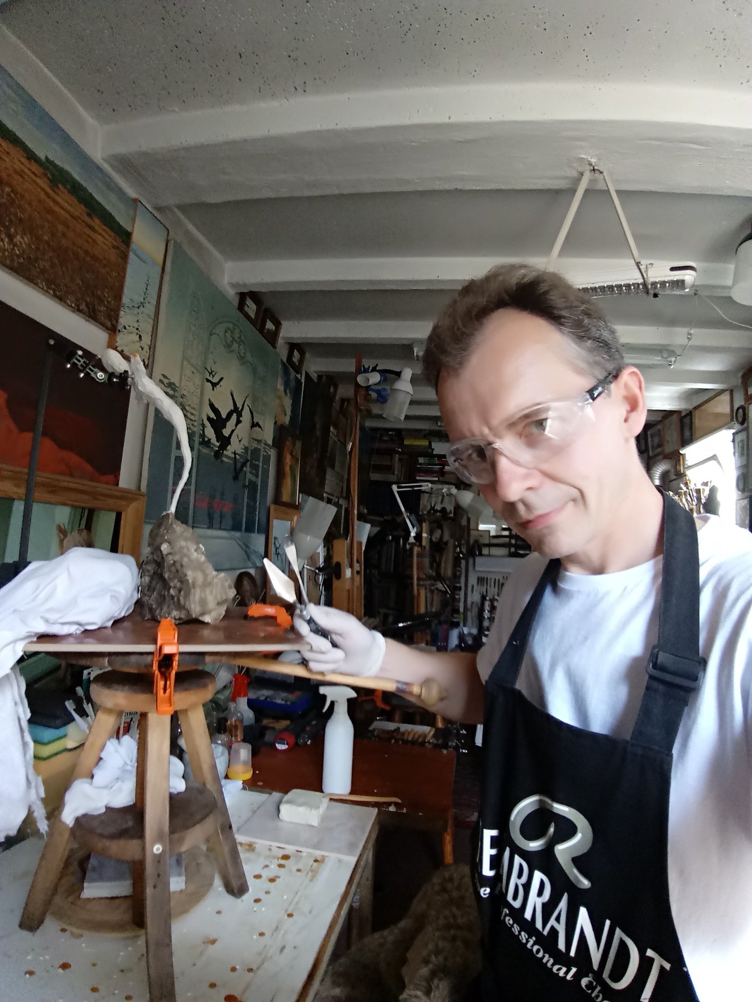 Artysta w okularach ochronnych i fartuchu z logo Rembrandt, trzymający szpachelki, pracuje nad rzeźbą ptaka na drewnianym stole w pracowni pełnej obrazów.