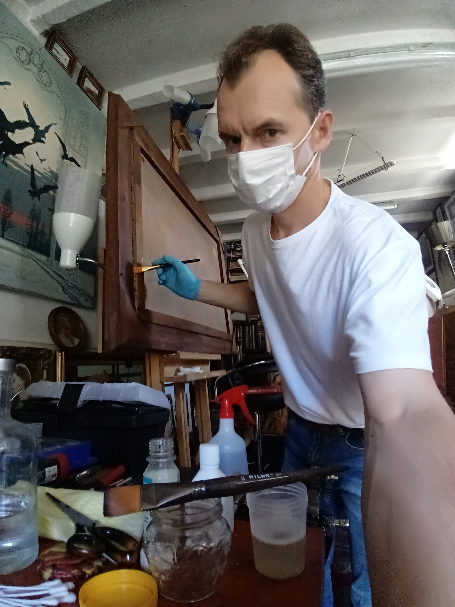 Mężczyzna w masce i rękawiczkach delikatnie odnawia ramę obrazu pędzlem w pracowni, na stole widoczne narzędzia i preparaty.