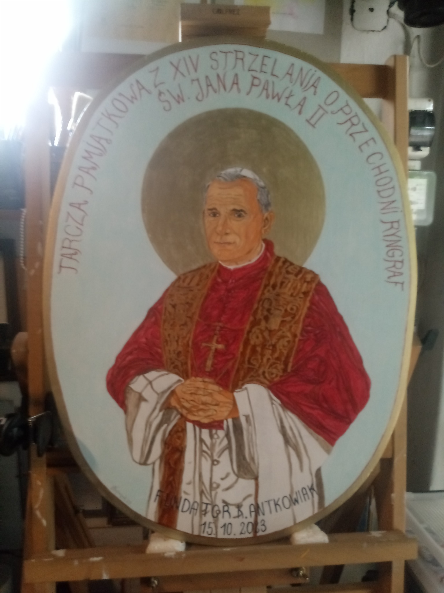 Owalna tarcza pamiątkowa z portretem św. Jana Pawła II, umieszczona na drewnianym stojaku, z inskrypcjami i datą 15.10.2013.