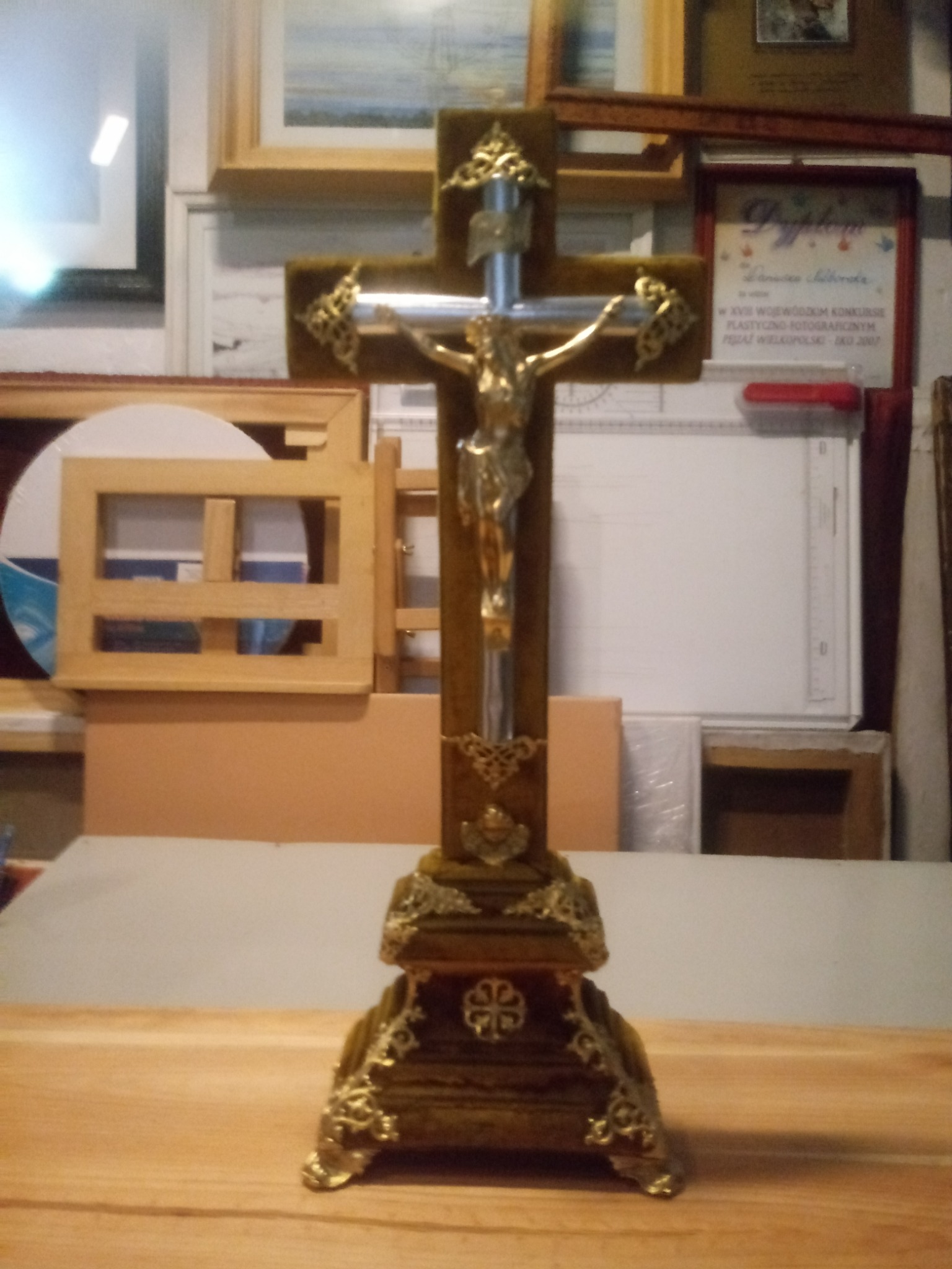 Drewniany, bogato zdobiony krucyfiks z metalową figurą Chrystusa na tle pracowni z ramami obrazów i dyplomem.
