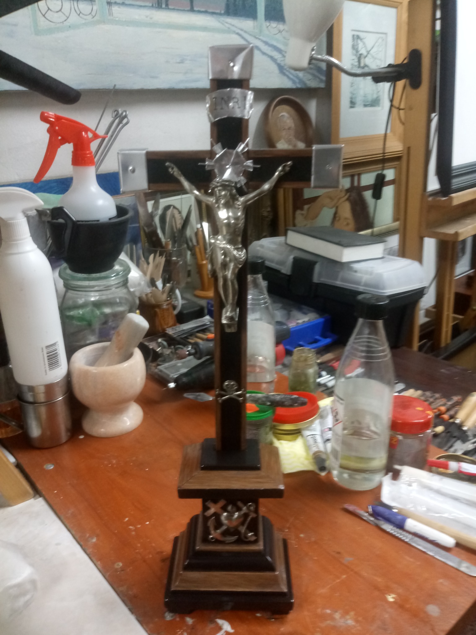 Krzyż z figurą Chrystusa na drewnianej podstawie, otoczony narzędziami i materiałami na stole warsztatowym, w tle obrazy i akcesoria malarskie.