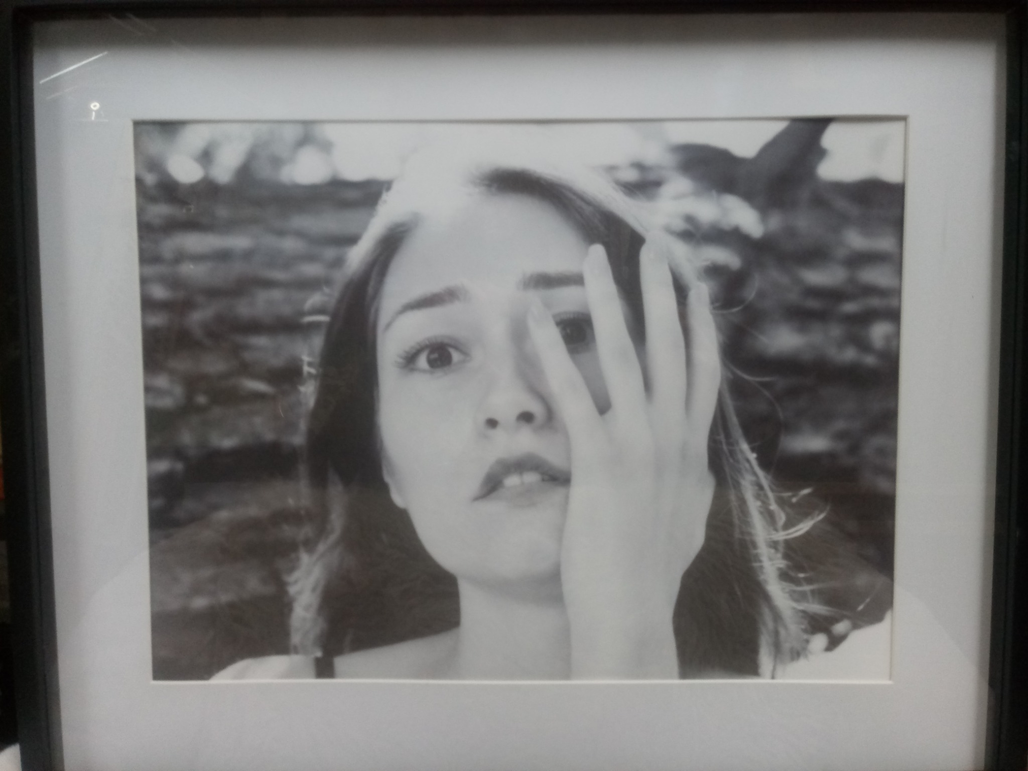 Oprawiona w ramę czarno-biała fotografia portretowa młodej kobiety, częściowo zasłaniającej twarz dłonią, z rozmytym tłem.