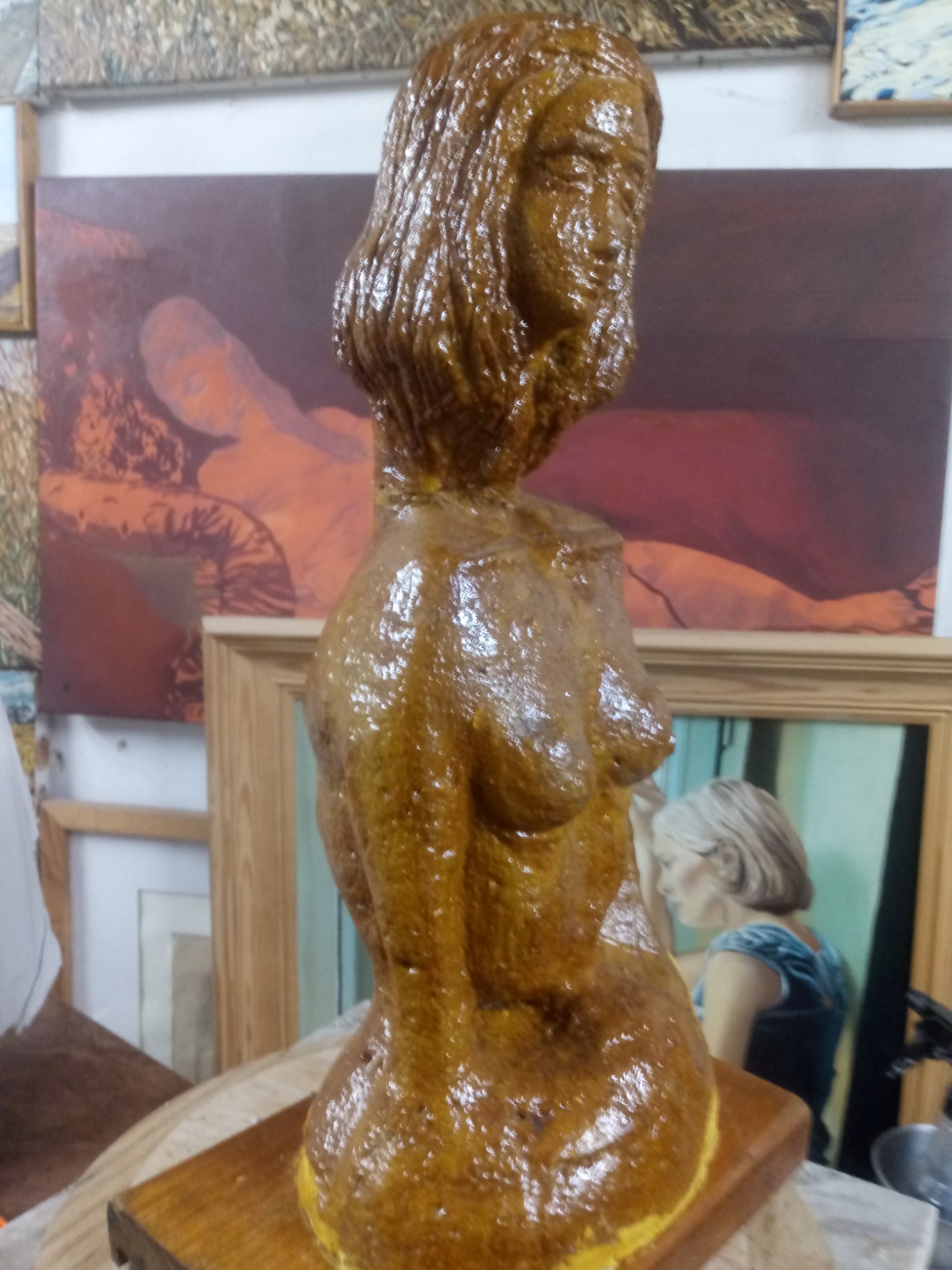 Bursztynowa, lakierowana rzeźba popiersia kobiety na drewnianym cokole, w tle obrazy w ramach, w tym akt kobiecy.