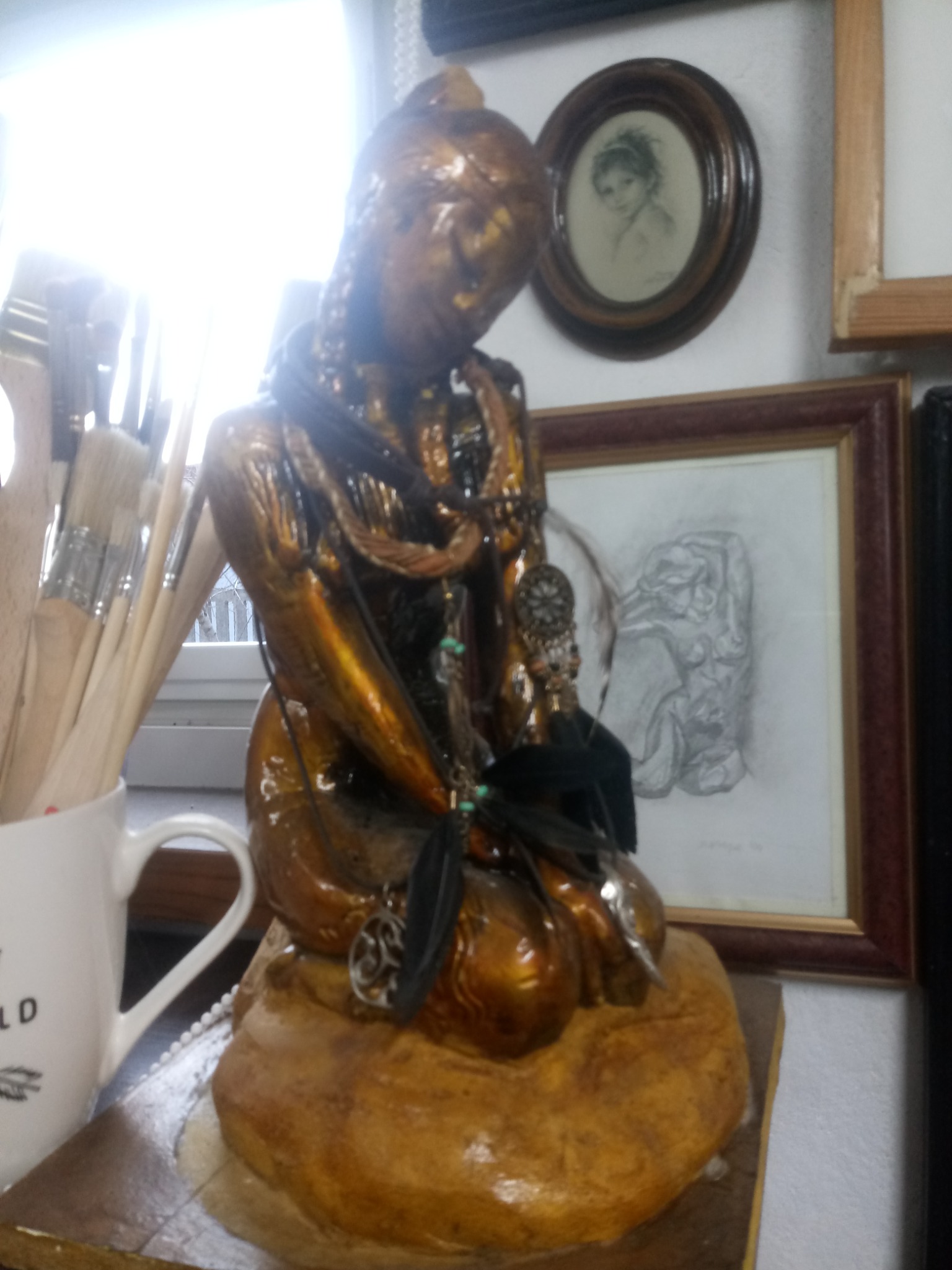 Złota, błyszcząca figurka kobiety z ozdobami, otoczona pędzlami w kubku, oprawionymi rysunkami i ramkami, ukazana w jasnym, naturalnym świetle.