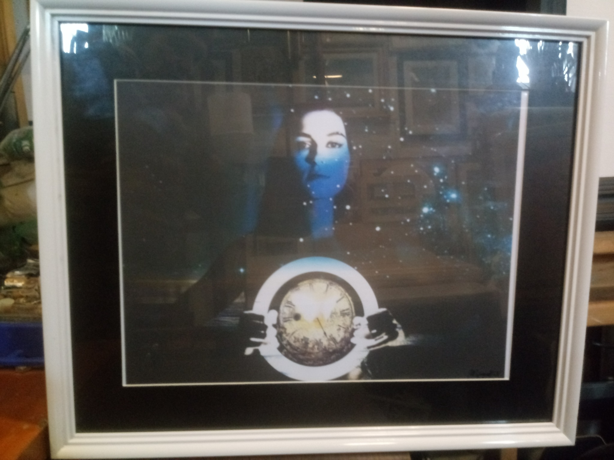 Oprawiony obraz przedstawiający postać z niebieską skórą, trzymającą zegar na tle kosmosu, w białej ramie z czarnym passe-partout.